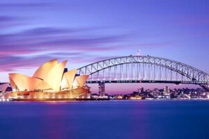 Sydney Harbour location showing the glowing Sydney Opera House and Sydney Harbour Bridge with lights against a beautiful magenta sunset. Business Electricity Prices can help your business reduce energy charges